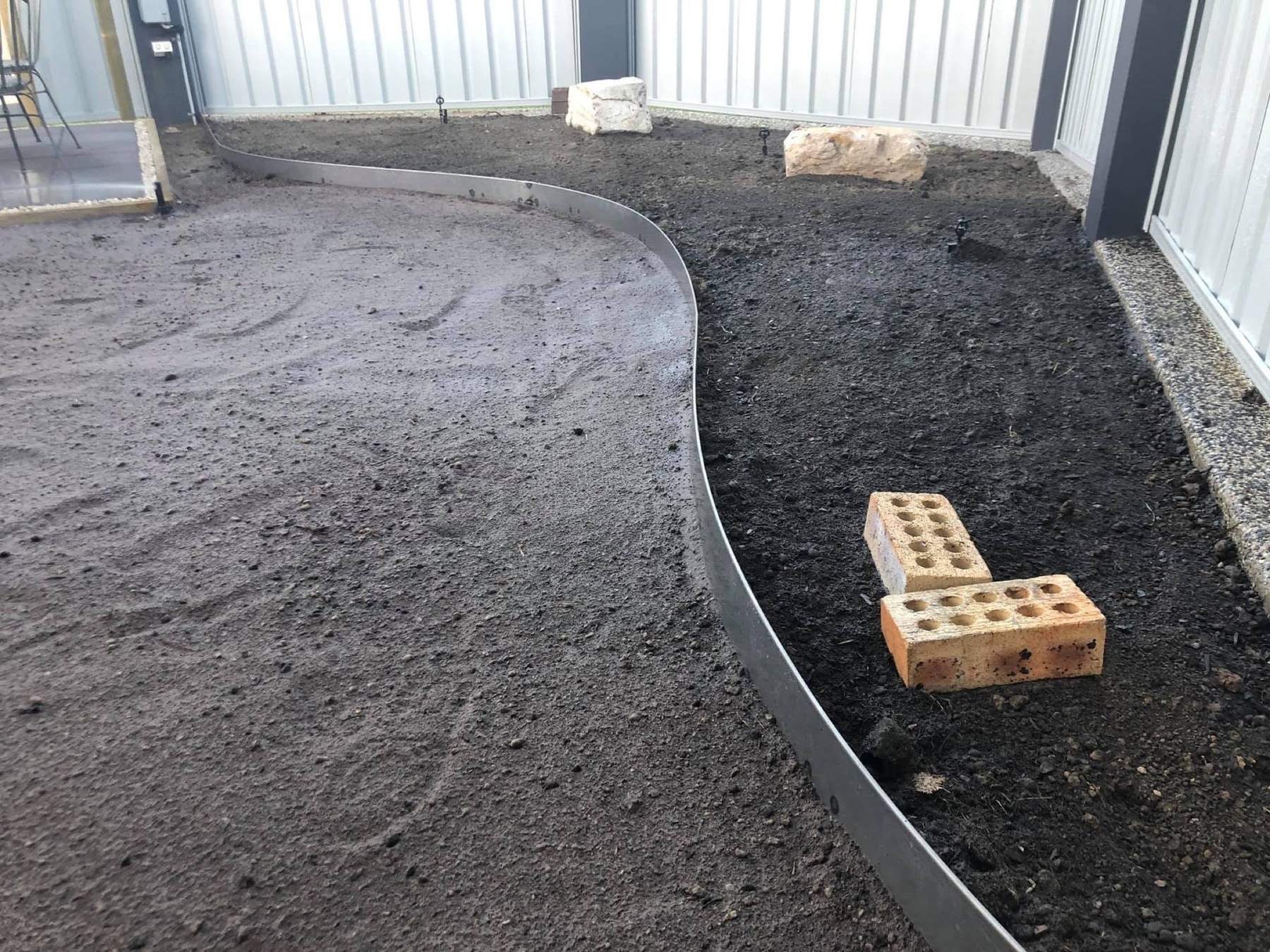 A steel garden edge curving through fresh soil beside a fence