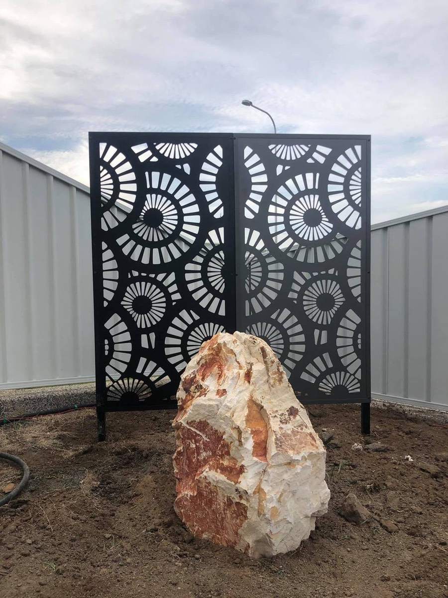 A dark laser-cut garden screen with a rock at its base against a fence