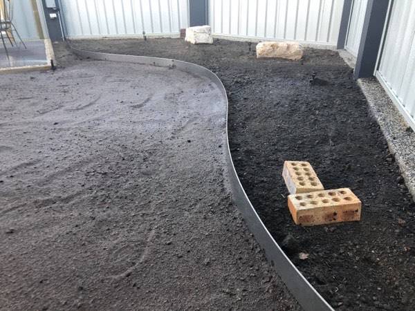 A steel garden edge curving through fresh soil beside a fence