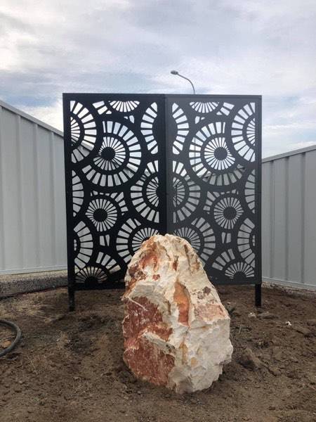 A dark laser-cut garden screen with a rock at its base